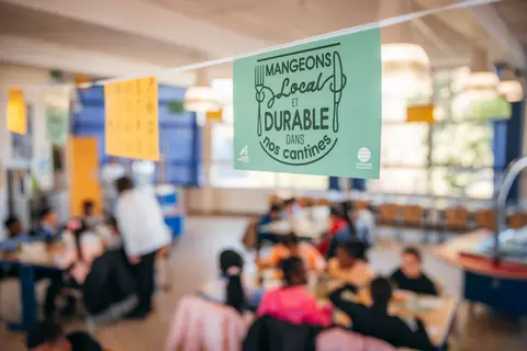Affiche "Mangeons local et durable dans nos cantines" avec en arrière plan des élèves dans un réfectoire